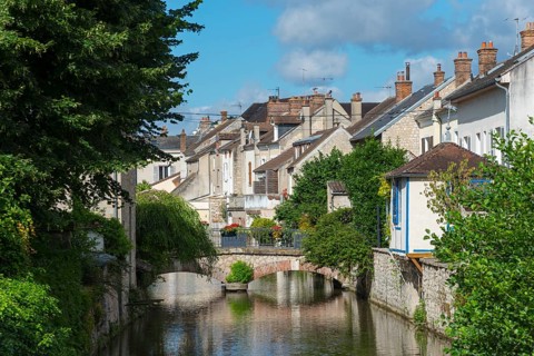 Malerisches Dorf mit Fluss, Brücke und dichten Häusern, umgeben von viel Grün und blauem Himmel,