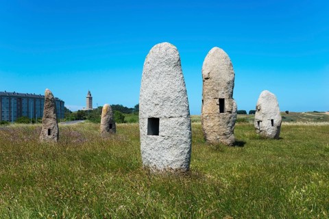 Stone formations in a meadow with a building in the background, under a clear blue sky, Menhires