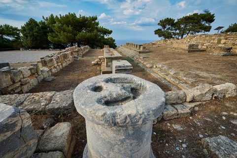 Ancient stone column with eroded details, surrounded by ruins and trees, Exedra, Kamiros,