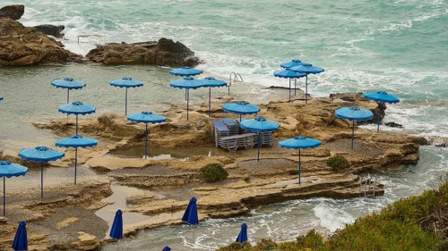 Rocks on the coast with blue parasols and deckchairs, surrounded by the sea with wavy water, Oasis