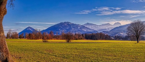 Green fields and trees in front of towering mountains under a clear blue sky, Bad Feilnbach