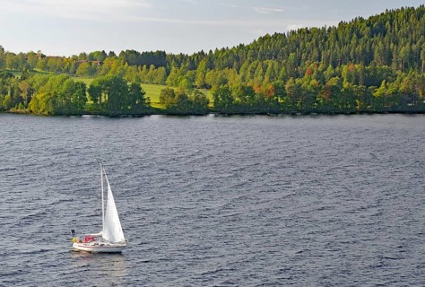 A sailboat sails on a calm lake surrounded by the green, wooded shores of Storsjon, Fröson Bridge,