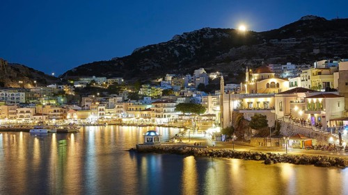 Illuminated coastal town at night with water and hills in the background, Pigadia, town and