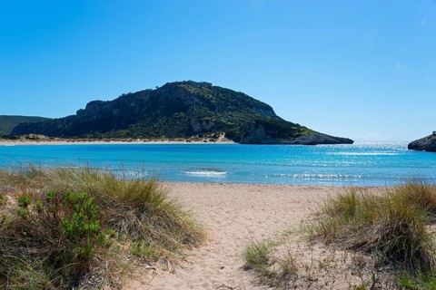 An idyllic beach with clear blue water and a mountain in the background on a sunny day, Voidokilia