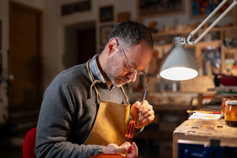 Violin maker working on a violin in his workshop, using a small brush and varnish, illuminated by a