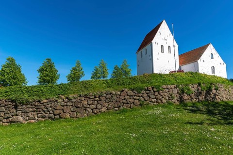 Ulbølle Kirke, whitewashed church on a hill, red roof tiles, group of trees, wall with boulders,
