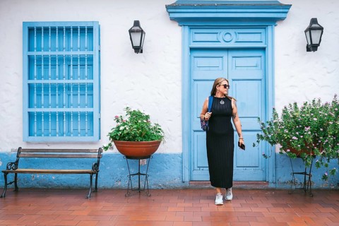 A fashionable woman in a black dress and sneakers walks past a blue colonial-style door and window