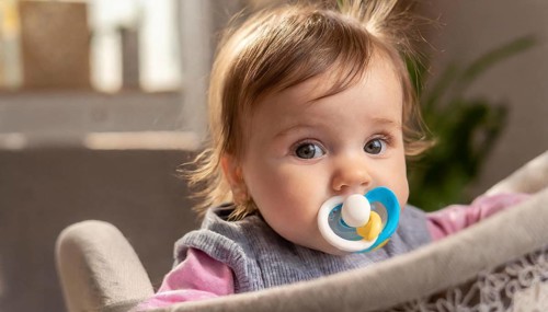 A baby with a blue dummy and blond hair sits in a cot and looks attentively, AI generated, AI