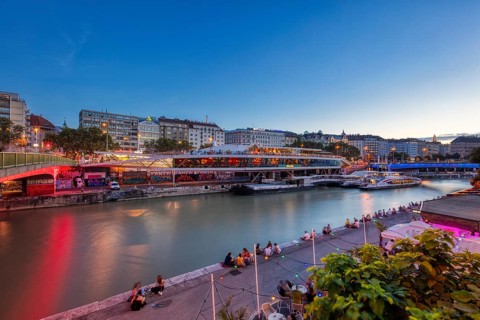 Evening scene by the river with illuminated promenade and people on the riverbank, relaxed