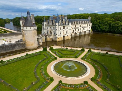 Castle and gardens with central fountain and geometric gardens by the river and under a cloudy sky,
