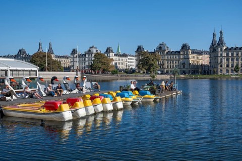 People enjoying leisure time on a jetty by the water, pedal boats, blue sky, historic buildings,