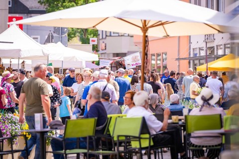 A lively market square in summer with many people sitting or walking under parasols, garden fair
