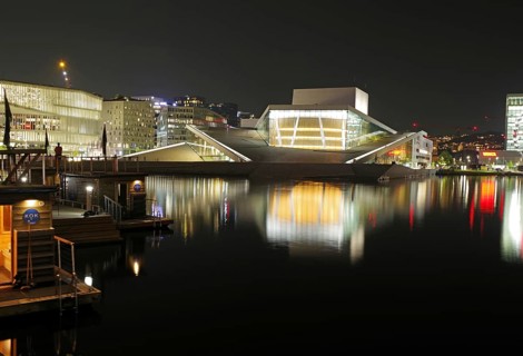 Illuminated opera house and modern buildings reflected in calm waters at night, Oslo city centre,
