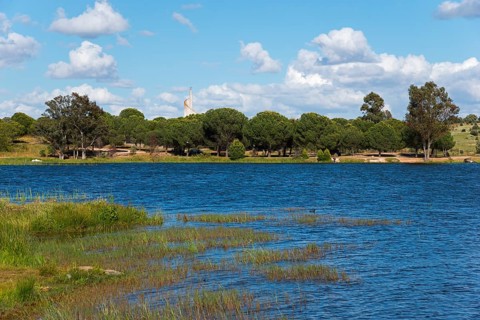 An idyllic lake with trees and blue water, framed by greenery and a sky with white clouds,