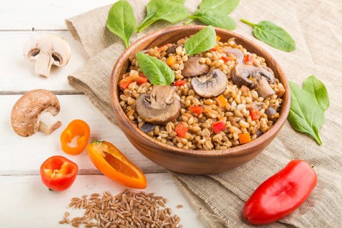 Spelt (dinkel wheat) porridge with vegetables and mushrooms in wooden bowl on a white wooden