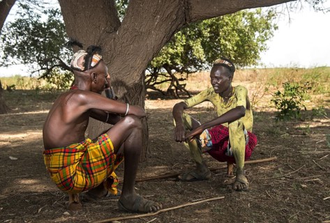 Two young men with traditional clay headdresses sitting on wooden headrests converted into stools,