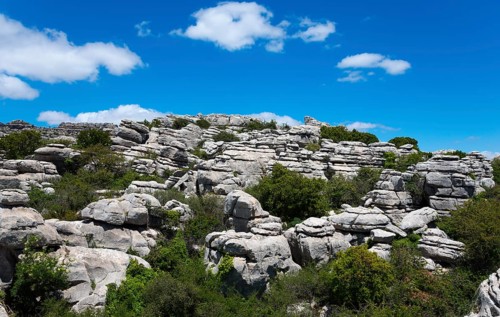 Barren rock formations under a bright blue sky with white clouds and green vegetation, karst