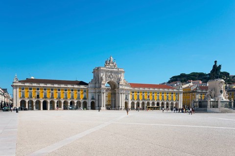 Spacious square with a large yellow building, arcades and a statue, all under a clear blue sky,