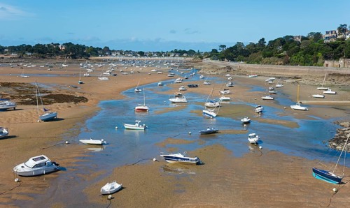 Many boats are scattered on the sandy shore at low tide, with the coast and houses behind them,