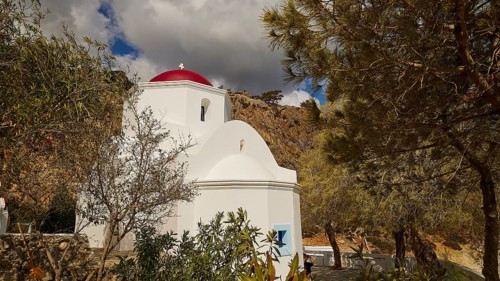 A picturesque white church with a red dome, surrounded by trees and rocks under a changeable sky,