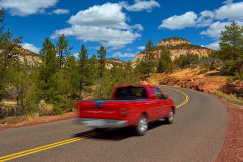 A red pickup truck drives on a winding road through a scenic mountain region with trees and blue