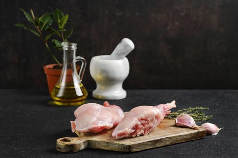 Fresh rabbit legs on a wooden board, accompanied by herbs, garlic, olive oil, and a mortar and