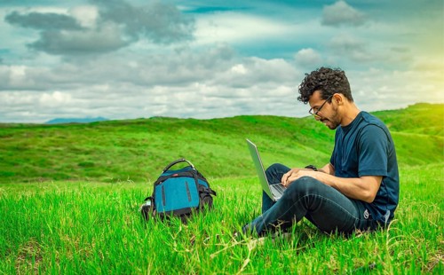 Smiling freelancer young man with laptop working from the field. Close up of man sitting on green