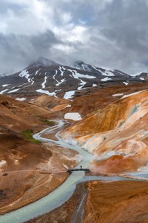 Tourist on bridge over steaming stream between colourful rhyolite mountains with snowfields,