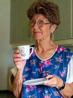 Granny with old smock apron, glasses and wig in the kitchen, holding coffee cup, Germany, Europe