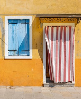 Yellow house facade with entrance door with red and white curtain and window with blue shutters,