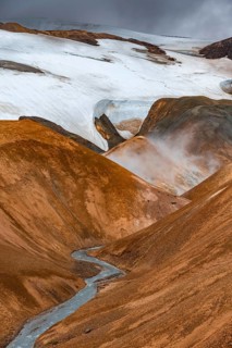 Steaming stream between colourful rhyolite mountains with snowfields, Hveradalir geothermal area,