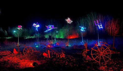 Artwork with colourful lights and chairs above wagon wheels at night, surrounded by trees, world of