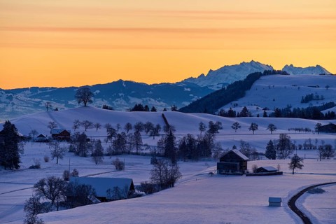 Snowy landscape at sunrise with mountains and farms, bathed in a warm orange-blue light, Menzingen,