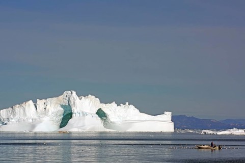 Small fishing boat surrounded by seabirds in front of iceberg with large ice cave, Ilulissat, Disko