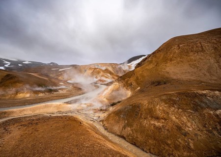 Steaming stream between colourful rhyolite mountains with snowfields, Hveradalir geothermal area,