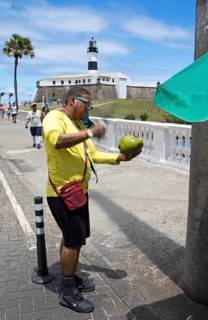 Brazilian man opening a coconut, Faro da Barra lighthouse in the back, Salvador, State of Bahia,
