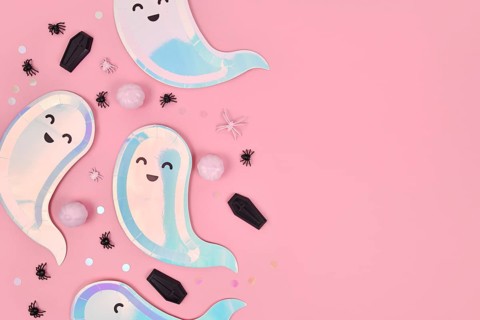 Cute pastel colored Halloween party flat lay with ghost shaped plates, coffins and confetti on pink