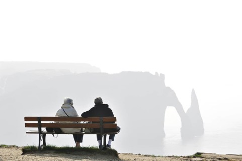 Elderly tourists sitting on bench looking at the Porte D'Aval, a natural arch in the chalk cliffs