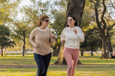 Two women run side by side in a lush green park on a sunny day. They enjoy the outdoors, staying