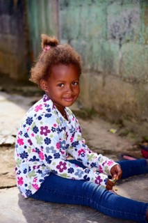 A smiling child with a floral print top sits casually in front of a house, between Pedro Sanchez
