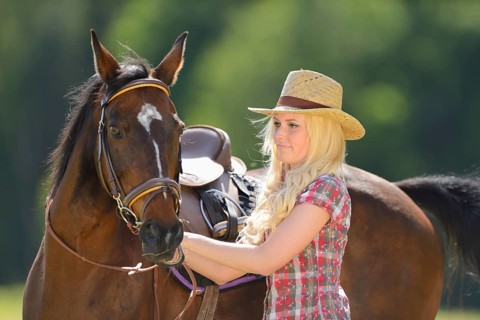 Woman (cowgirl) in a straw hat and plaid shirt stands beside a polish arabian horse on a field,