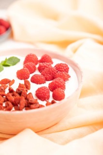 Yogurt with raspberry and goji berries in ceramic bowl on white concrete background and orange
