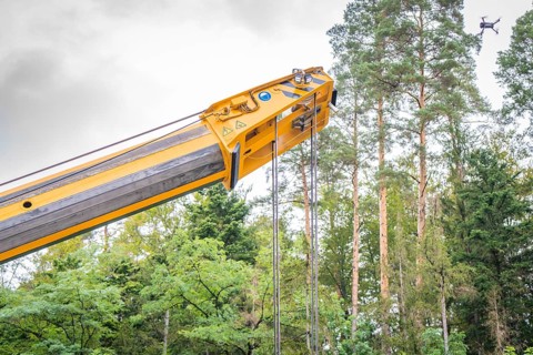 Yellow crane arm with ropes and a flying drone in front of tall trees in the forest, track