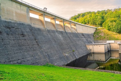 An imposing concrete dam with flowing water, embedded in a green hilly landscape,