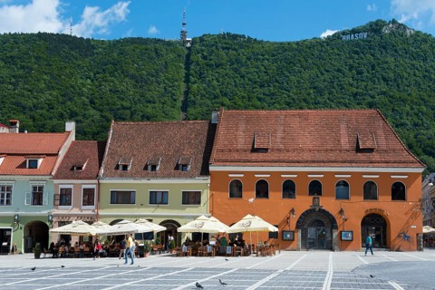A lively town square in front of historic buildings with cafés at the foot of a green mountain,