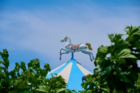 A carousel horse stands on a blue and white striped roof, surrounded by green leaves under a clear