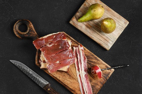 Cured duck meat on a wooden board next to two ripe pears. A knife rests nearby, highlighting the