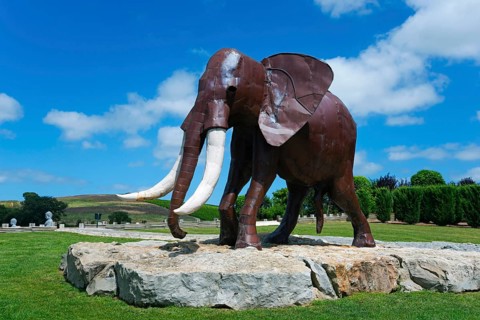 Metal sculpture of an elephant on a rock foundation in a green park under a blue sky with white