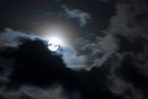 The full moon shines brightly in a cloudy, stormy, black sky and has a halo (corona),