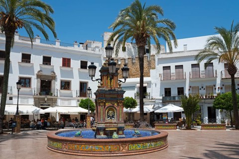 A central fountain in a sunny plaza surrounded by white buildings and palm trees, fountain with